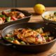 Three chicken dinners with vegetables, pasta, lemons, garlic, and herbs on a rustic table.