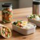 A bento box surrounded by a wrap, salad jar, eggs, fruit, and thermos.
