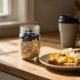 A jar of blueberry oats sits beside eggs, yogurt, toast, and a travel mug.