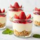 Glass jars layered with strawberry cheesecake, crumb topping, berries, and mint on marble.